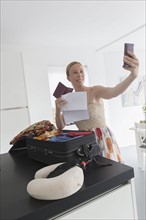 Happy mature woman packing suitcase for vacations and taking selfie