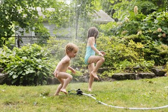 Naked boy and his sister playing with sprinkler