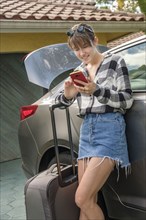 Woman with smart phone and suitcase by car