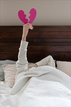 Woman holding aloft pink broken heart while lying in bed