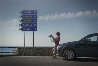 Woman holding map leaning on car by road sign