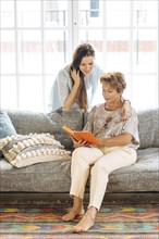 Mother and adult daughter looking at book on sofa