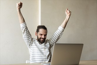 Smiling young businessman with his arms raised by laptop