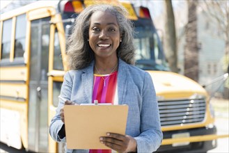 Smiling teacher with clipboard by school bus