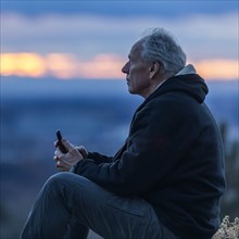 Seated man holding smart phone at sunset