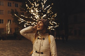 Smiling woman wearing coat by fairy lights in tree at night