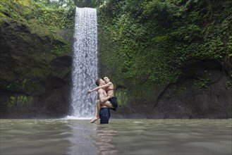 Young man carrying young woman in river by Tibumana Waterfall in Bali, Indonesia