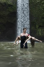 Young man carrying young woman in river by Tibumana Waterfall in Bali, Indonesia