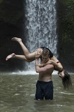 Young man carrying young woman over his shoulder in river by Tibumana Waterfall in Bali, Indonesia