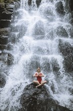 Woman wearing red swimsuit sitting by waterfall in Bali, Indonesia