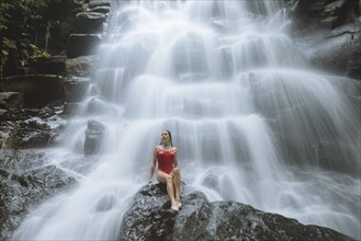 Woman wearing red swimsuit sitting by waterfall in Bali, Indonesia