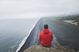 Man wearing red coat sitting above beach in Vik, Iceland