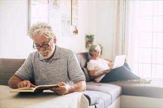 Senior man reading book as his wife uses laptop in living room