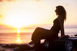 Woman sitting on car by beach at sunset