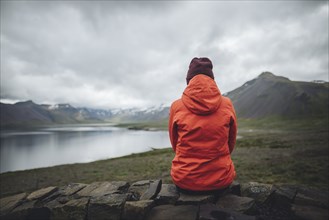 Woman wearing red raincoat sitting by fjord in Snufellsnes, Iceland