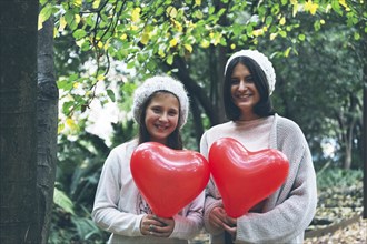 Smiling girls holding heart balloon by trees
