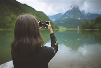 young woman photographing mountain and lake with smartphone