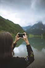 young woman photographing mountain and lake with smartphone