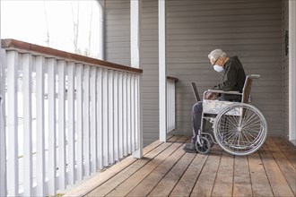Senior man in wheelchair wearing protective mask to prevent coronavirus transmission using laptop on porch
