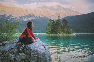 Germany, Bavaria, Eibsee, Young woman sitting on rock and looking at scenic view by Eibsee lake in Bavarian Alps