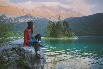 Germany, Bavaria, Eibsee, Young woman sitting on rock and looking at scenic view by Eibsee lake in Bavarian Alps
