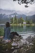 Germany, Bavaria, Eibsee, Young woman sitting on rock at shore of Eibsee lake in Bavarian Alps