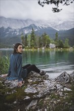 Germany, Bavaria, Eibsee, Portrait of young woman sitting on rock at shore of Eibsee lake in Bavarian Alps