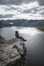 Norway, Senja, Man taking photo sitting on edge of steep cliff on top of mountain Segla