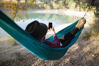 Italy, Man lying in hammock near lake and using smart phone