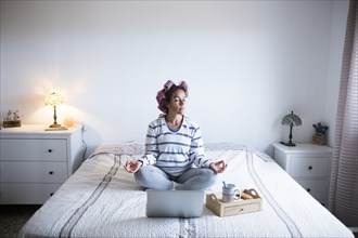 Woman with hair curlers meditating on bed in front of laptop