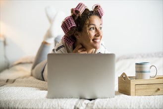 Woman with hair curlers lying on bed with laptop