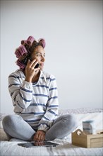 Woman with hair curlers sitting on bed and talking on mobile phone