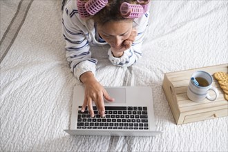 Woman with hair curlers lying on bed and using laptop