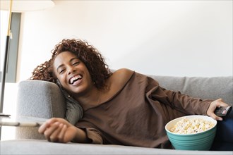 Laughing woman on sofa with TV remote and popcorn