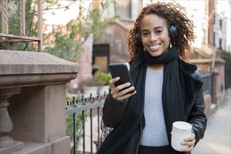 Smiling woman wearing headphones holding smart phone and cup