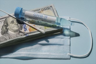 Covid-19 test vial and swab on stack of 100 dollar bills and medical mask