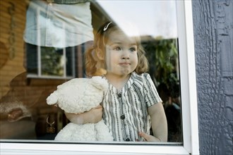 Girl (2-3) holding stuffed toy and looking through window