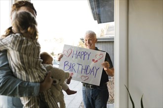 Grandfather showing happy birthday message to family with grandchildren (2-3 months, 2-3) through window