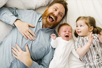 Laughing father lying on bed with crying baby boy (2-3 months) and daughter (2-3)