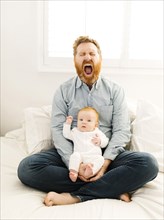Yawning father sitting on bed with baby boy (2-3 months)