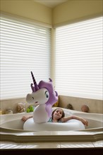 Girl (6-7) in unicorn shaped pool float in bathtub
