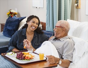 Daughter visiting senior father eating healthy meal in bed in nursing home