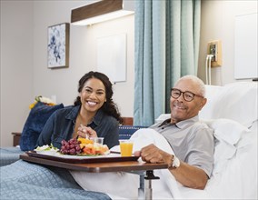 Daughter visiting senior father eating healthy meal in bed in nursing home