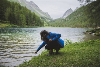 Switzerland, Bravuogn, Palpuognasee, Young woman crouching by Palpuognasee lake in Swiss Alps