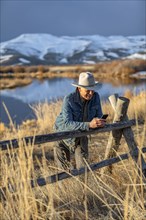 USA, Idaho, Sun Valley, Senior man in cowboy hat leaning against fence texting