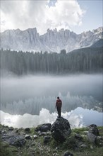 Italy, Carezza, Young man standing on rock at Lago di Carezza in Dolomite Alps at dawn