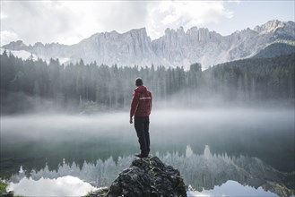 Italy, Carezza, Young man standing on rock and looking at Lago di Carezza in Dolomite Alps at dawn