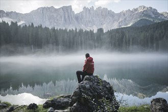 Switzerland, Young man sitting at Lago di Carezza in Dolomite Alps at dawn