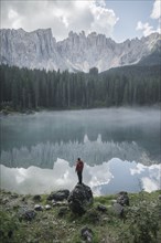 Italy, Carezza, Young man standing on rock at Lago di Carezza in Dolomite Alps at dawn