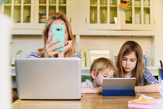 Woman holding smart phone in front of laptop while boy (4-5) and girl (6-7) using digital tablet